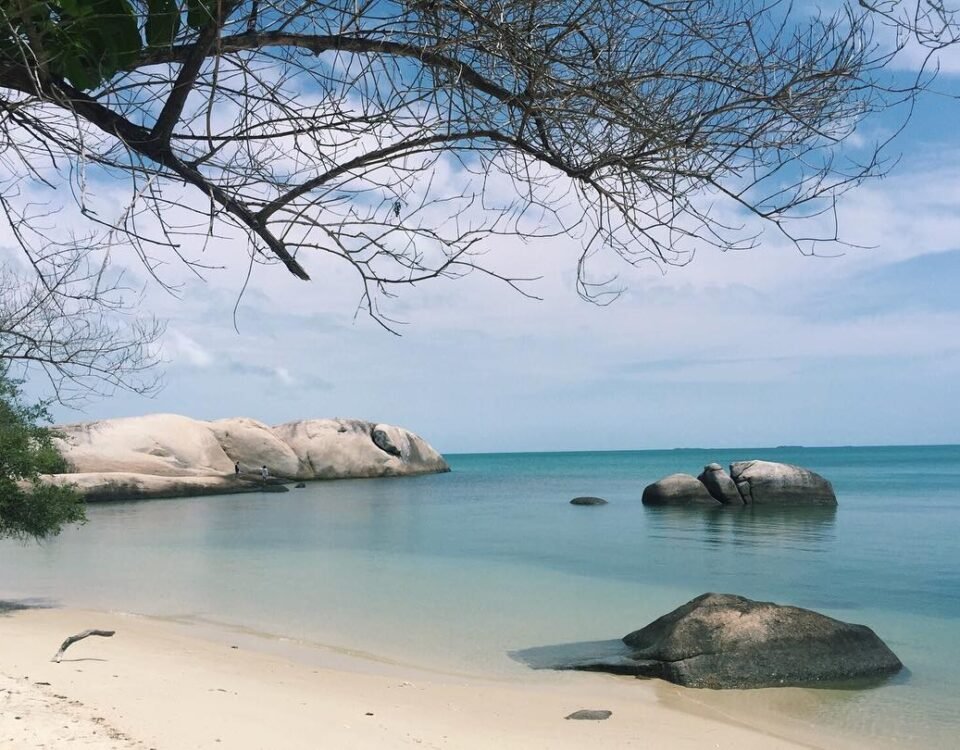 Peaceful tropical beach with pale sand, calm turquoise water, and large granite rocks along the shoreline under a blue sky.