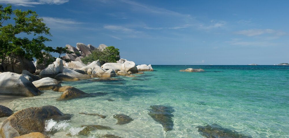 Clear turquoise water along a rocky tropical beach with green trees on the left and a blue sky above.