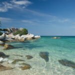 Clear turquoise water along a rocky tropical beach with green trees on the left and a blue sky above.