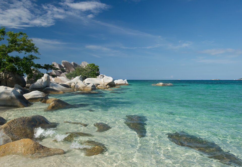 Clear turquoise water along a rocky tropical beach with green trees on the left and a blue sky above.