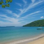 Sunny tropical beach with clear turquoise water, white sandy shore, a small boat near the shoreline, and green hills in the background.