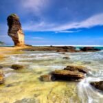 Tall sea stack rises from a rocky shore under a clear blue sky with shallow tidal pools and waves around flat rocks on the coast.