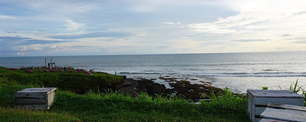 Green grassy foreground with wooden crates overlooking a rocky shoreline and calm ocean under a cloudy sky at sunset; people picnicking on the left edge.