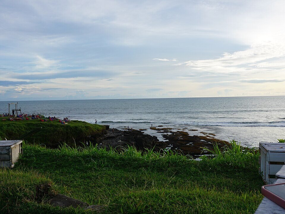 Green grassy foreground with wooden crates overlooking a rocky shoreline and calm ocean under a cloudy sky at sunset; people picnicking on the left edge.