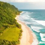 A sandy beach runs along a lush green cliffside with waves crashing on shore on a sunny day.