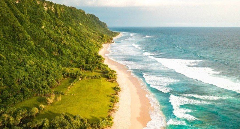A sandy beach runs along a lush green cliffside with waves crashing on shore on a sunny day.