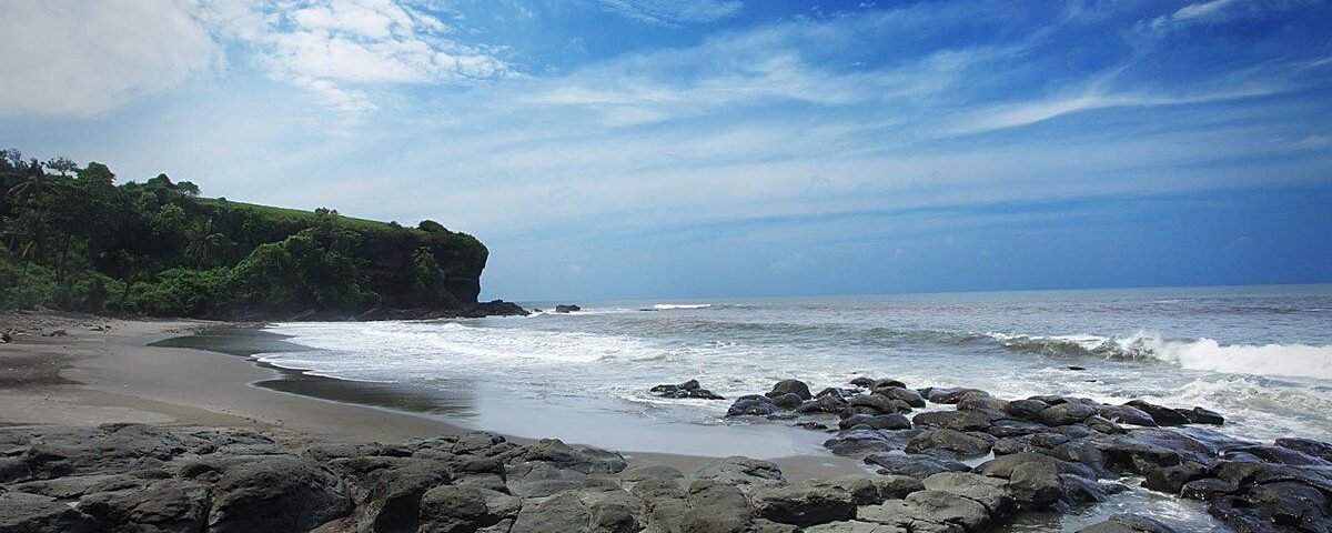 Rocky shoreline and sandy beach with a green cliff on the left under a blue sky and scattered clouds, waves at the shore.