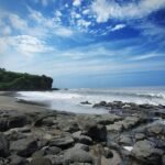 Rocky shoreline and sandy beach with a green cliff on the left under a blue sky and scattered clouds, waves at the shore.