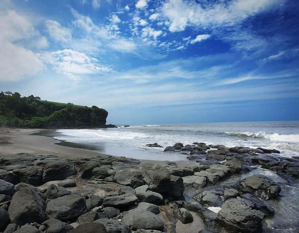 Rocky shoreline and sandy beach with a green cliff on the left under a blue sky and scattered clouds, waves at the shore.
