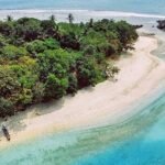 Aerial view of a tropical island with dense green jungle, white sandy beach, and turquoise water along the shore.