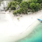 A small tropical beach with white sand, clear turquoise water, and dense green trees; a blue boat rests near the shoreline.