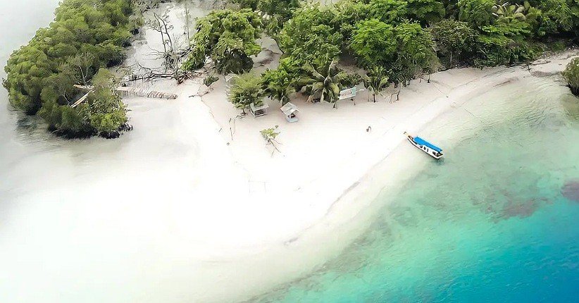 A small tropical beach with white sand, clear turquoise water, and dense green trees; a blue boat rests near the shoreline.
