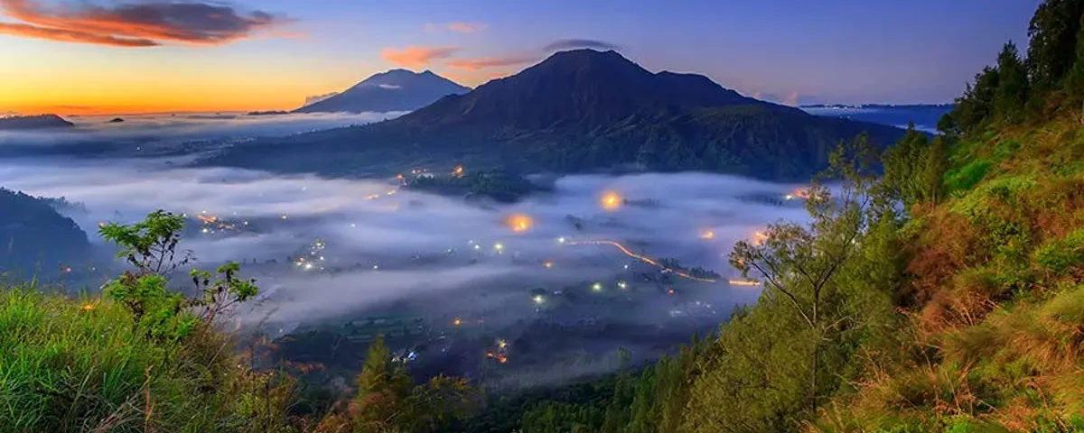 Sunrise over a mountain range with a fog-filled valley and twinkling village lights below, surrounded by green hills and trees in the foreground.