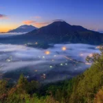 Sunrise over a mountain range with a fog-filled valley and twinkling village lights below, surrounded by green hills and trees in the foreground.
