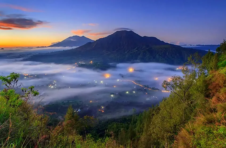 Sunrise over a mountain range with a fog-filled valley and twinkling village lights below, surrounded by green hills and trees in the foreground.