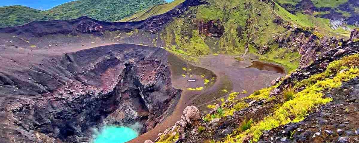 Volcanic crater lake with turquoise water, surrounded by dark lava rocks and green hills.