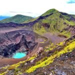 Volcanic crater lake with turquoise water, surrounded by dark lava rocks and green hills.