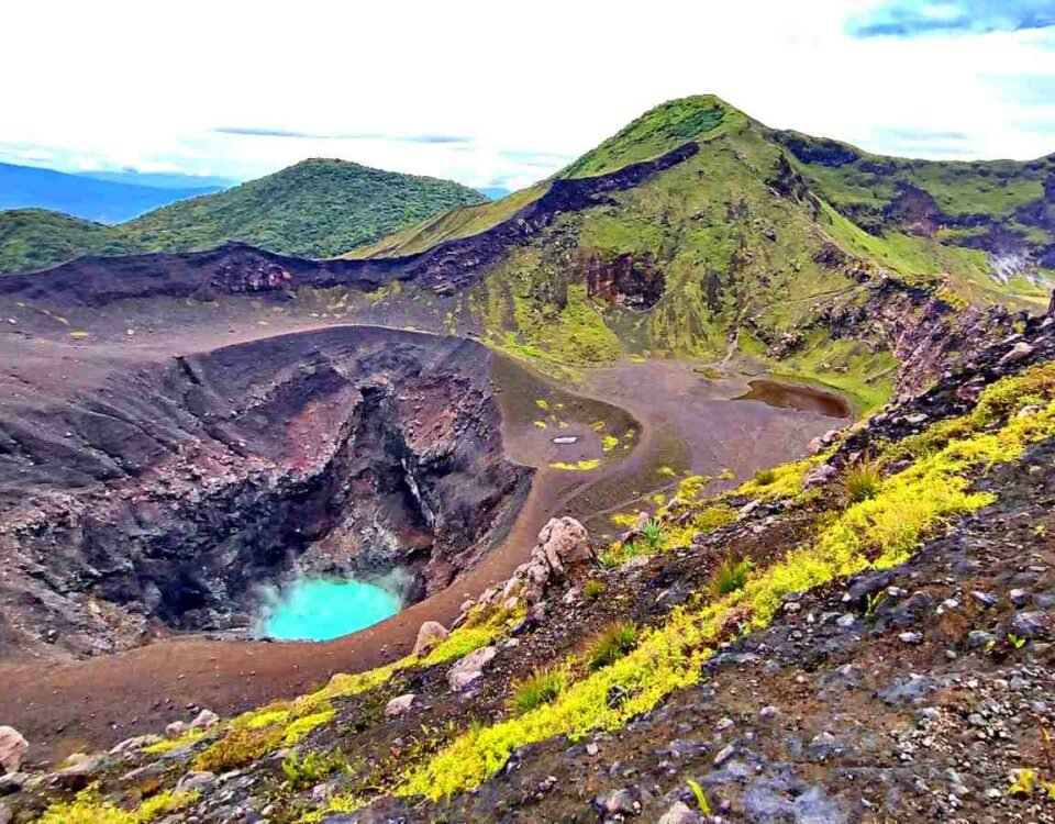 Volcanic crater lake with turquoise water, surrounded by dark lava rocks and green hills.