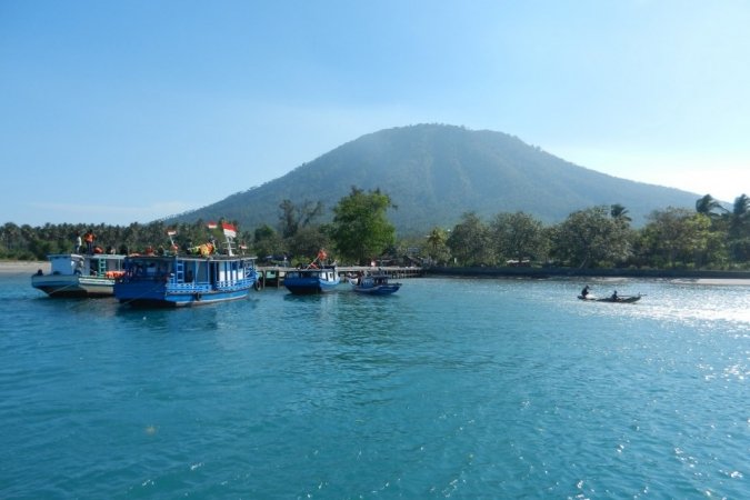 Blue passenger boats anchored near a tropical shoreline with a forested mountain in the distance.