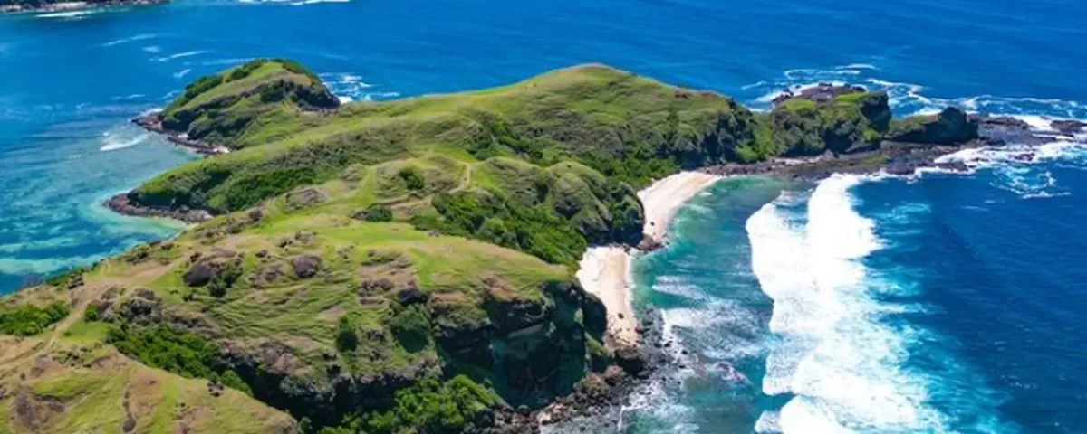 Aerial view of green cliffs and a sandy cove along a turquoise sea.