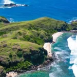 Aerial view of green cliffs and a sandy cove along a turquoise sea.