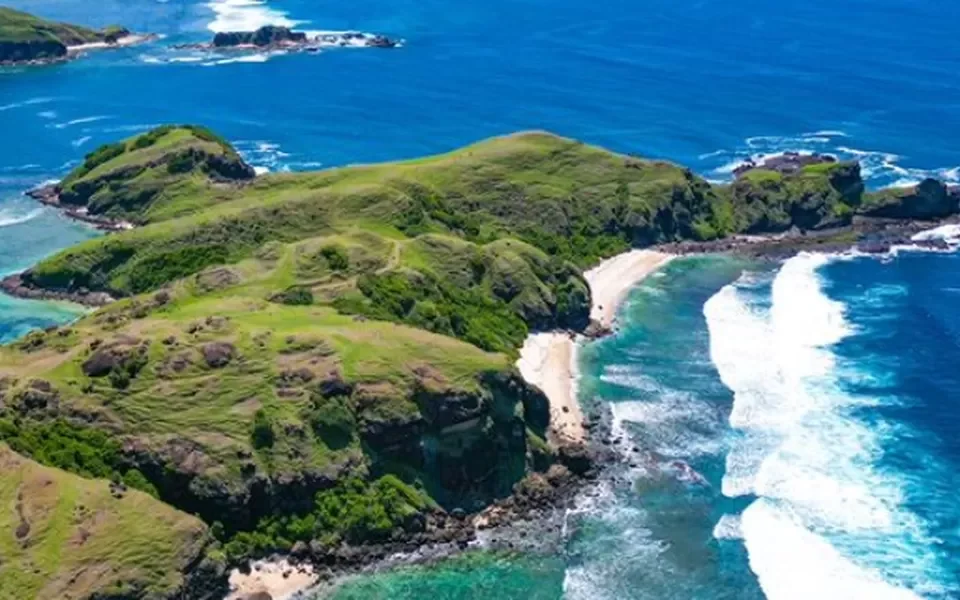 Aerial view of green cliffs and a sandy cove along a turquoise sea.