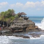 People walk along a rocky seaside outcrop with a thatched-roof pavilion overlooking the water and waves crashing nearby.