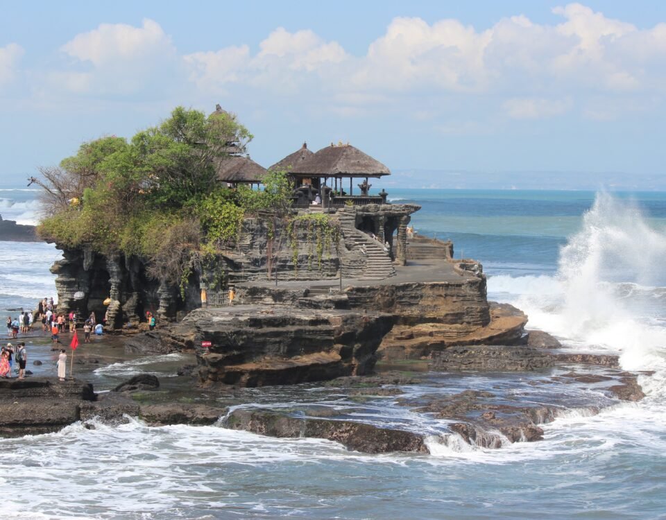 People walk along a rocky seaside outcrop with a thatched-roof pavilion overlooking the water and waves crashing nearby.