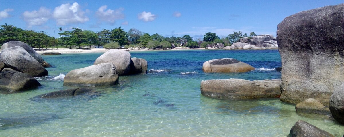Clear turquoise water lapping a sandy beach with large granite boulders and green trees under a bright blue sky.