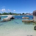 Clear turquoise water lapping a sandy beach with large granite boulders and green trees under a bright blue sky.