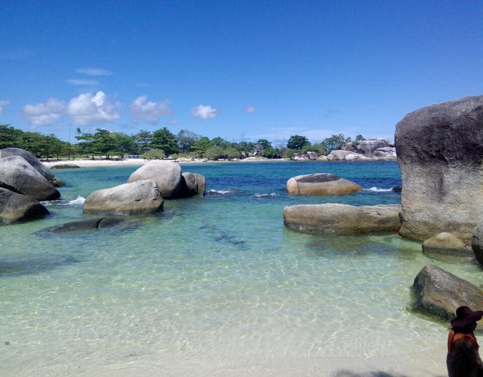 Clear turquoise water lapping a sandy beach with large granite boulders and green trees under a bright blue sky.