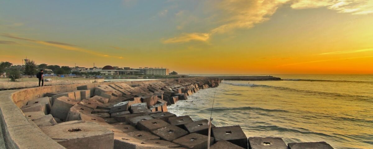 Sunset over a breakwater lined with concrete tetrapods; a lone walker along the promenade by the calm sea.