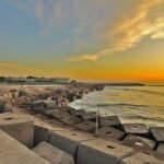 Sunset over a breakwater lined with concrete tetrapods; a lone walker along the promenade by the calm sea.