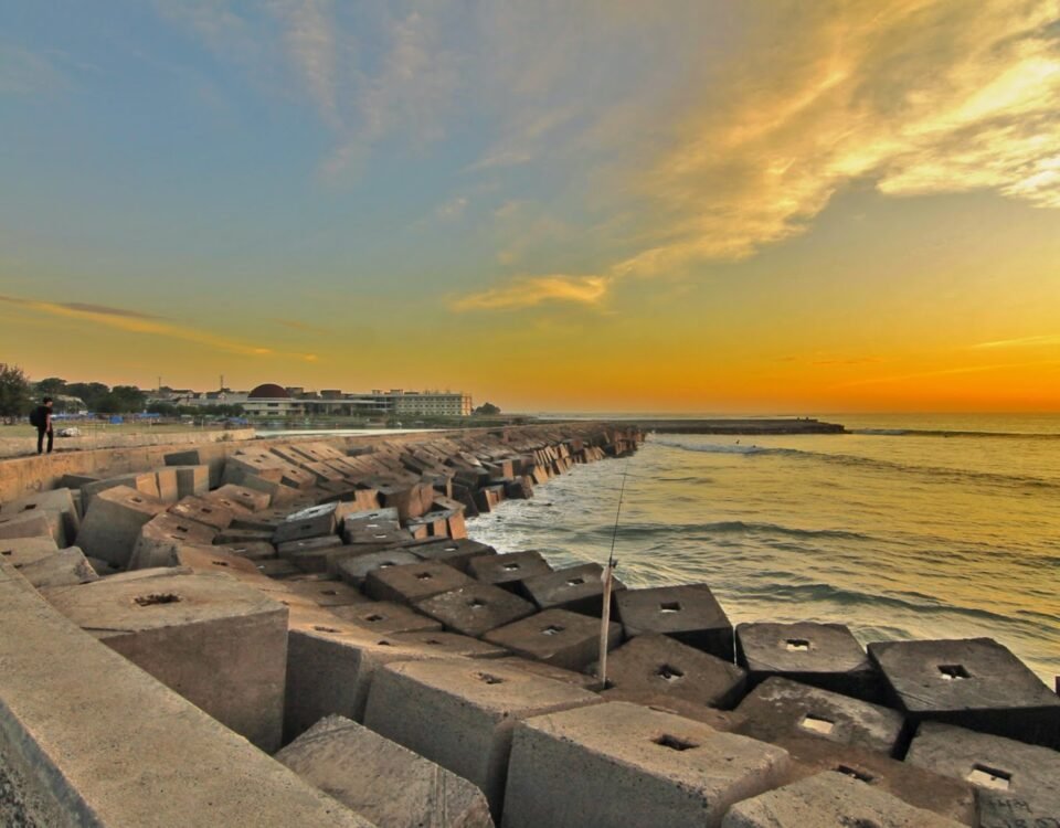 Sunset over a breakwater lined with concrete tetrapods; a lone walker along the promenade by the calm sea.