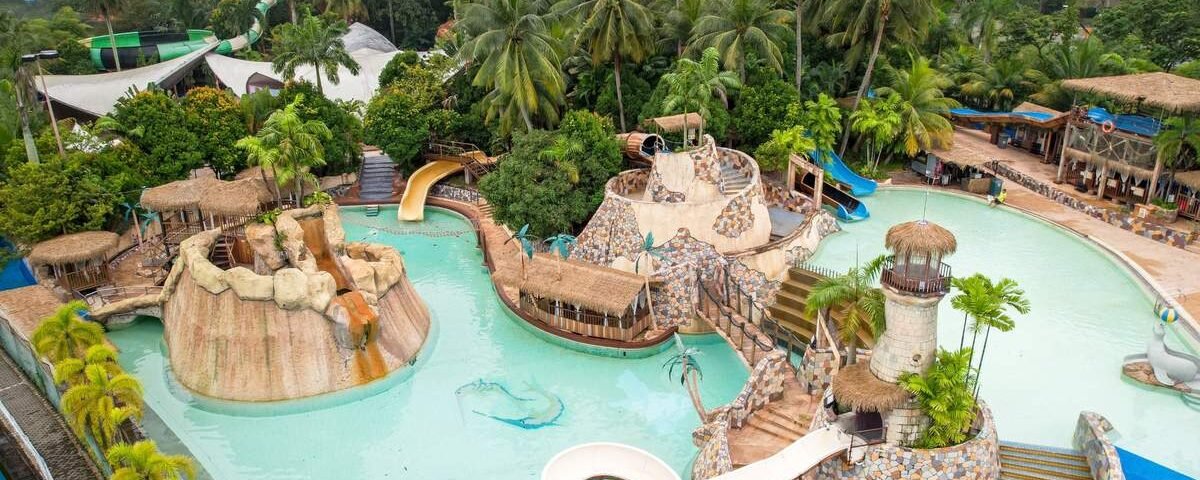 Aerial view of a tropical water park with turquoise water, winding slides, rock formations, and palm trees.