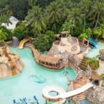 Aerial view of a tropical water park with turquoise water, winding slides, rock formations, and palm trees.