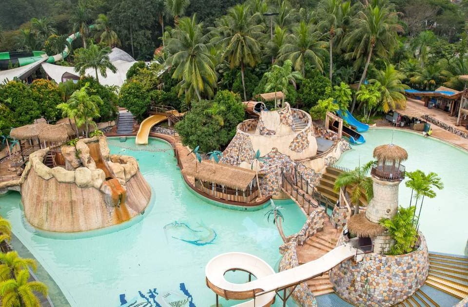 Aerial view of a tropical water park with turquoise water, winding slides, rock formations, and palm trees.