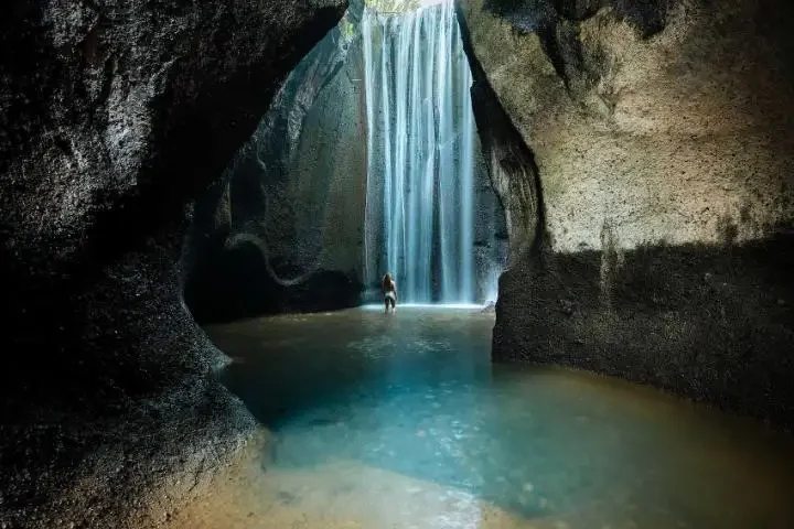 Cave with a tall waterfall pouring into a turquoise pool, a person standing waist-deep in the water.