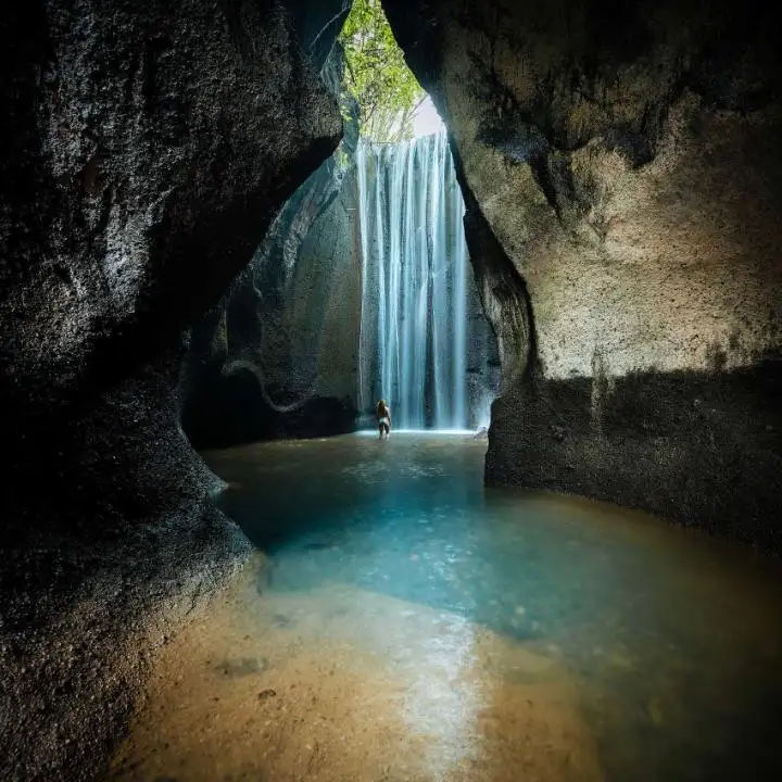 Cave with a tall waterfall pouring into a turquoise pool, a person standing waist-deep in the water.
