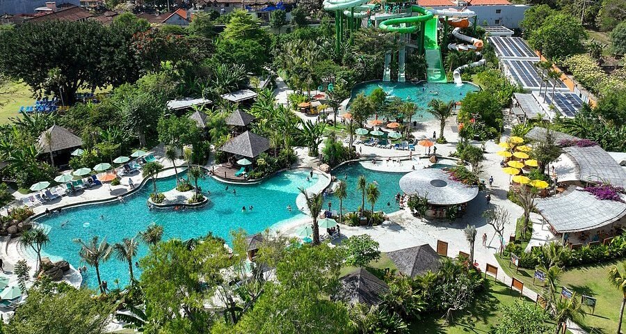 Aerial view of a resort water park with turquoise pools, twisting green slides, and palm trees scattered around.