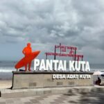 Orange surfer statue beside a large 'PANTAI KUTA' sign on a stone pedestal at the beach, with the sea and cloudy sky in the background.