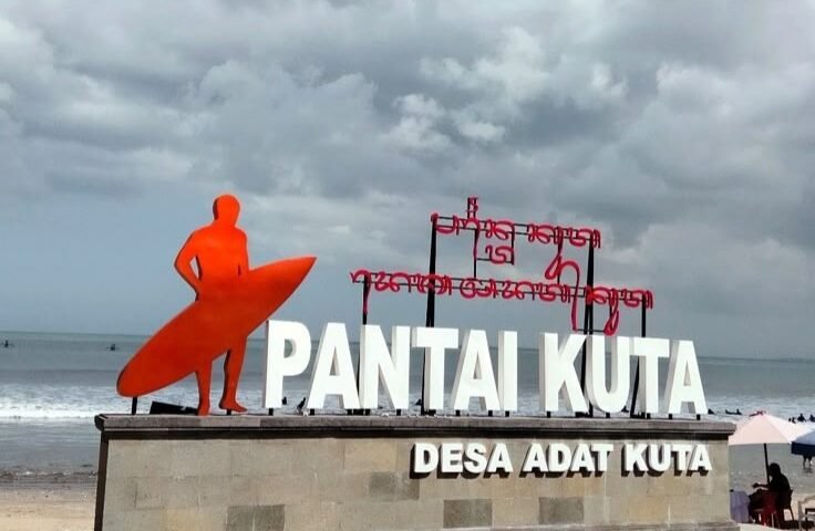 Orange surfer statue beside a large 'PANTAI KUTA' sign on a stone pedestal at the beach, with the sea and cloudy sky in the background.