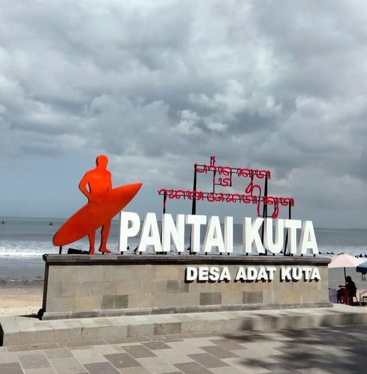 Orange surfer statue beside a large 'PANTAI KUTA' sign on a stone pedestal at the beach, with the sea and cloudy sky in the background.