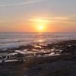 Sunset over the calm ocean with a rocky shoreline and tide pools in the foreground.