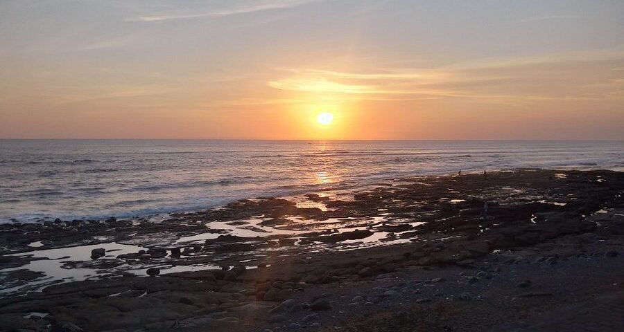 Sunset over the calm ocean with a rocky shoreline and tide pools in the foreground.