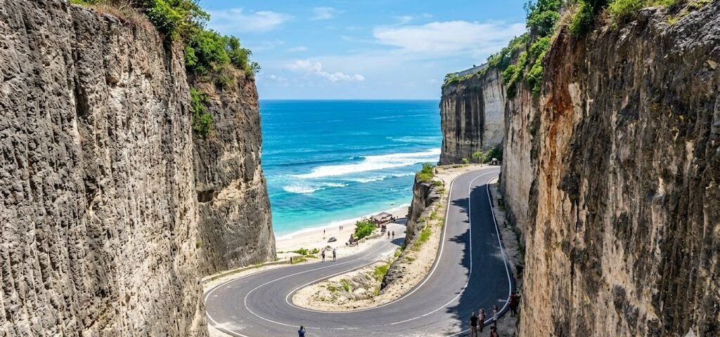 Winding coastal road between tall rocky cliffs, leading to a sandy beach and blue sea beneath a clear sky.