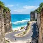 Winding coastal road between tall rocky cliffs, leading to a sandy beach and blue sea beneath a clear sky.
