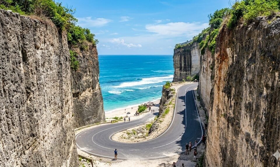 Winding coastal road between tall rocky cliffs, leading to a sandy beach and blue sea beneath a clear sky.
