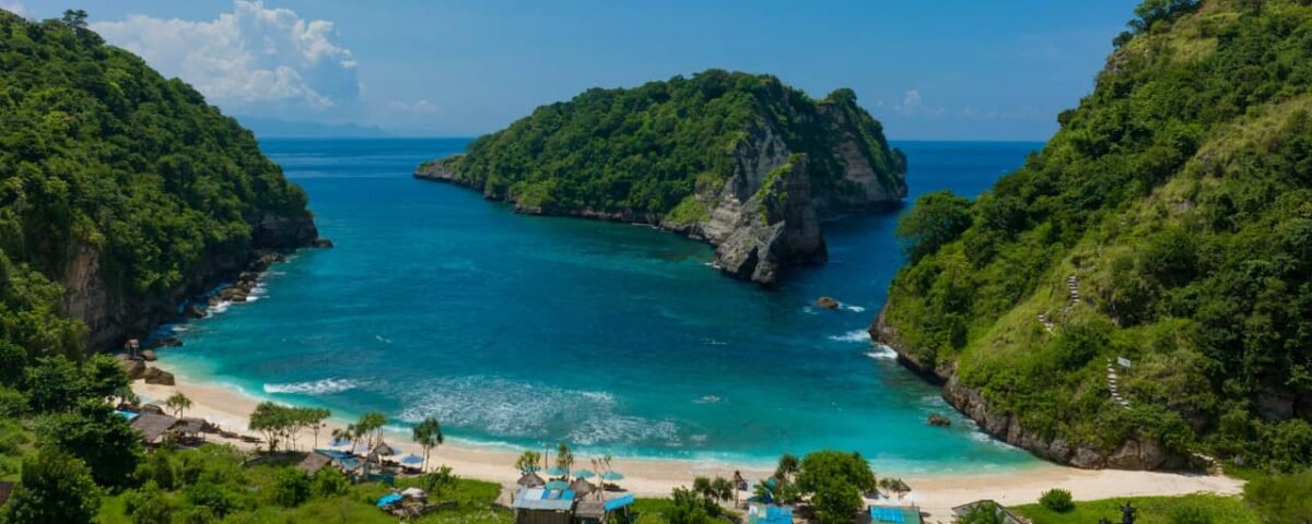 Tropical cove with turquoise water, white sand beach, and lush green cliffs; small beach huts line the shore.