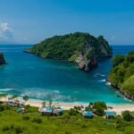 Tropical cove with turquoise water, white sand beach, and lush green cliffs; small beach huts line the shore.